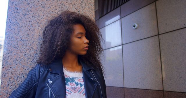 A teenage girl with curly hair and a leather jacket is standing outside a modern, tiled building. She appears confident and focused. This can be used for themes about youth fashion, confidence, urban lifestyle, or modern architecture.