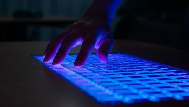Hovering hand over backlit rgb keyboard emitting blue purple neon glow on wooden desk