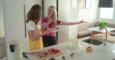 Two women decorating cake, playfully arguing in bright modern kitchen with frosting, fruit