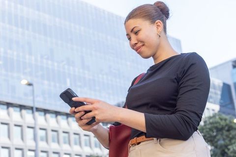 African businesswoman standing in city using smartphone and card reader with red shoulder bag