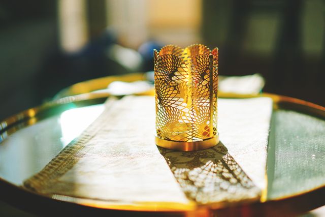 Golden metal candle holder sitting on glass table, intricately carved pattern casting beautiful shadows in sunlight. Ideal for use in home decor projects, interior design inspiration content, and light study backgrounds.