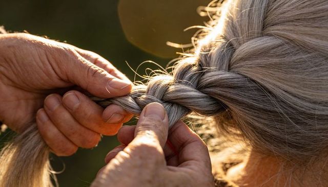 Hands braiding silver-gray hair into three-strand braid during warm golden hour light