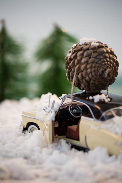 Toy car transporting a large pine cone on snow with green trees in the background. Ideal for holiday greeting cards, Christmas-themed advertisements, festive home décor ideas, or blog posts about holiday crafts and decorations.