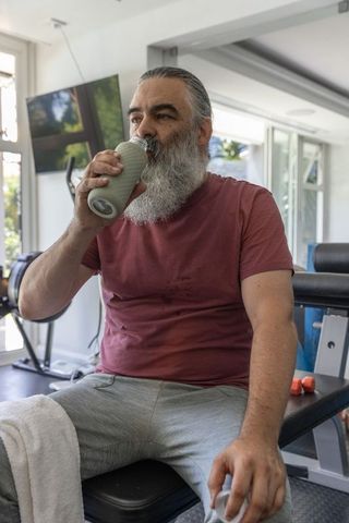 Mature man sipping from textured reusable bottle while resting on bench in home gym