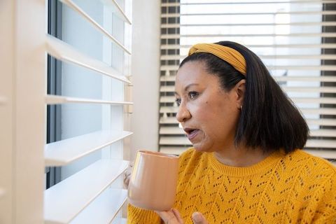 Mature woman gazing through blinds holding mug in yellow sweater, quiet morning