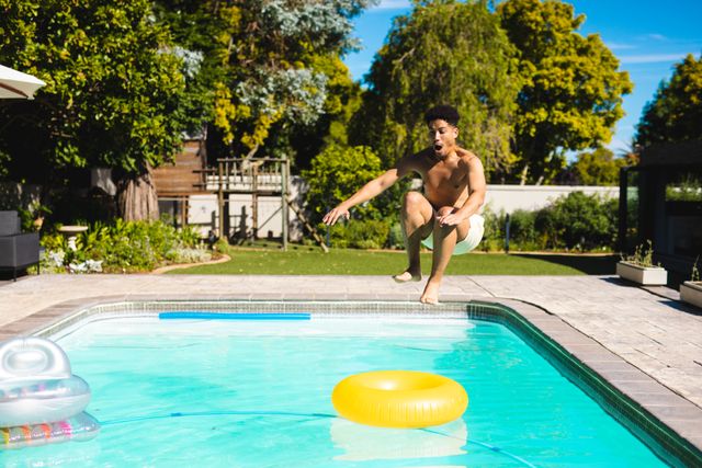 Shirtless Hispanic man mid-air jumping into a backyard swimming pool on a sunny day. Inflatable toys float on the water. Ideal for use in summer activity promotions, leisure and vacation advertisements, and lifestyle blogs focusing on outdoor fun and relaxation.