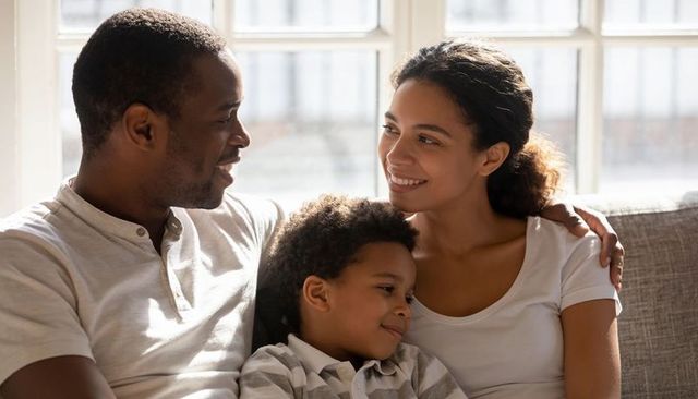 Smiling family cuddling on sofa, parents embracing child by sunlit multi-pane window