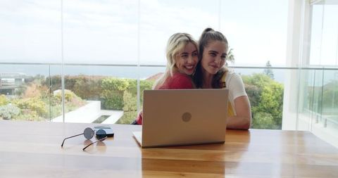 Two friends hugging while working on laptop by glass balcony overlooking ocean
