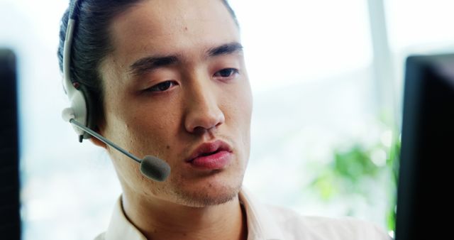 An Asian young man is focused on his work as a customer service representative, wearing a headset with a microphone, with copy space. His professional demeanor suggests he is attentively handling a customer's query or issue.