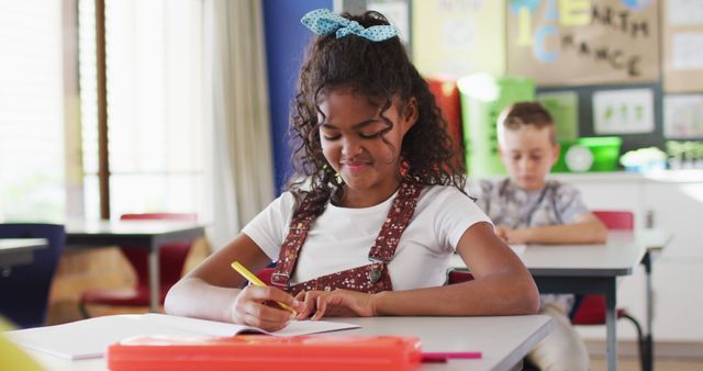 Portrait of happy biracial schoolgirl sitting at classroom, making notes, looking at camera. children at primary school