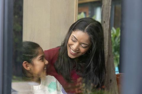 Diverse mother and daughter smiling and sharing moment by window in cozy knit sweaters
