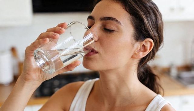 Young woman drinking water from clear glass in kitchen wearing white tank top, hydration