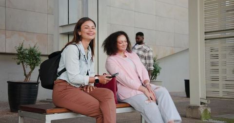Two friends relaxing on bench in urban plaza holding smartphone and backpacks