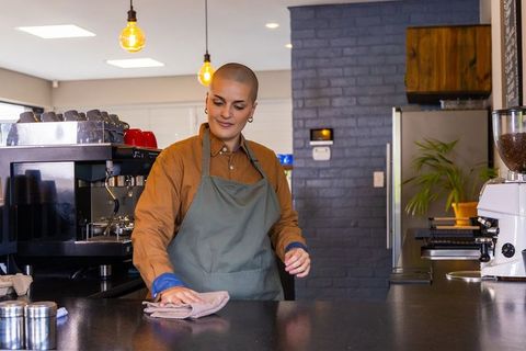 Female barista wiping counter in modern coffee shop wearing green apron, warm lighting