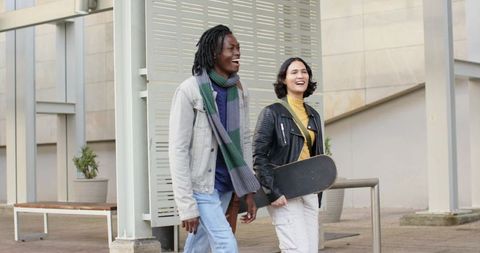 Diverse friends laughing and walking through modern urban plaza carrying skateboard
