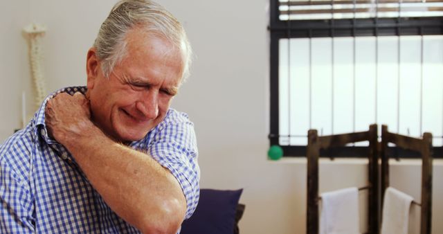 A senior Caucasian man appears to be in discomfort as he holds his shoulder, indicating pain or injury. His expression suggests he may be experiencing a moment of distress or physical discomfort related to his shoulder.