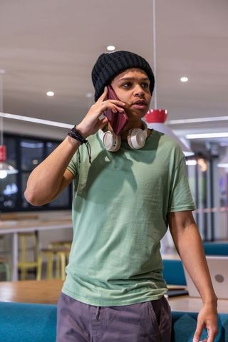 College student talking on phone in campus lounge wearing beanie and headphones