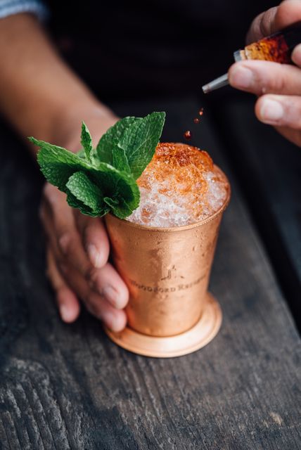 Hand holding copper mug filled with iced cocktail. Mint leaves and bitters added on top as garnish. Perfect for promoting cocktail recipes, refreshing summer drinks, or craft beverage preparation tutorials. Ideal for bars, restaurants, or cocktail-making blogs.