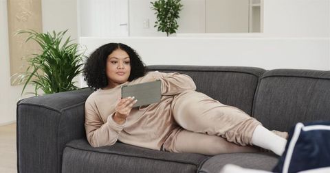 Midlife African American woman relaxing on modern sofa in beige loungewear holding tablet