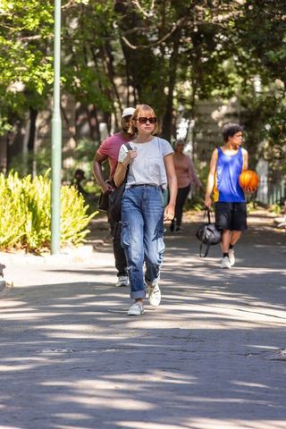 Woman walking in sunlit park wearing sunglasses and carrying shoulder bag, urban pedestrians