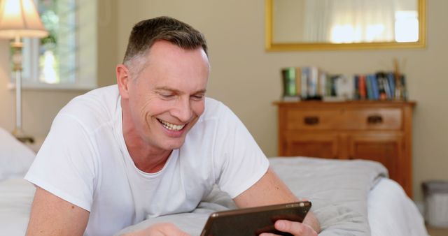 Middle-aged man lying on bed and using tablet in bedroom. Ideal for concepts such as home relaxation, casual daily life, modern technology use, leisure time, and comfortable living environments.