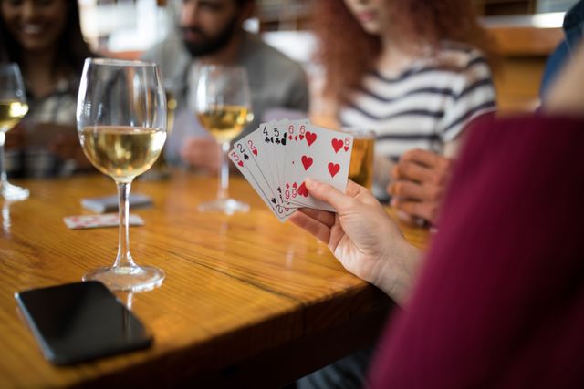 Group of friends enjoying a casual evening playing cards in a bar. Wine glasses and a smartphone are on the table, indicating a relaxed and social atmosphere. Ideal for use in articles or advertisements about social gatherings, leisure activities, or bar promotions.