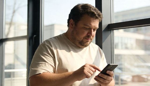 Adult man using smartphone at modern office window in daylight, urban mobile lifestyle