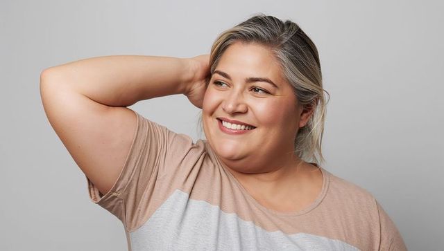 Confident plus-size woman smiling while raising arm behind head in casual neutral portrait