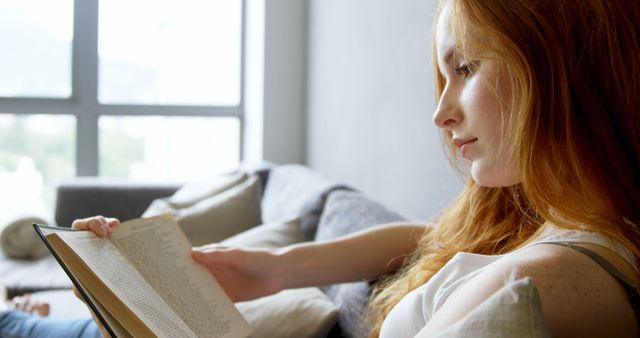 Young Caucasian woman enjoys reading at home, with copy space. She's immersed in a book, highlighting a moment of leisure and learning.