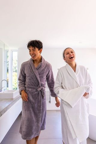 Diverse couple laughing in modern bathroom wearing plush bathrobes