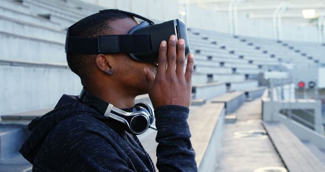 Man wearing VR headset engaging with virtual reality outdoors, large seating area in background. Great for illustrating modern technology, innovations in digital experiences, or use in tech-centric advertisements and promotional materials.