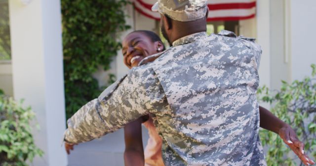 African american male soldier hugging smiling wife in front of house with american flag. soldier returning home to family.