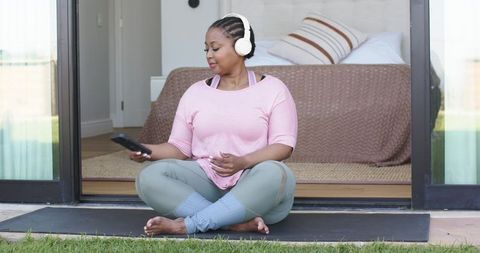 Mid-adult african american woman relaxing on yoga mat with headphones at bedroom threshold