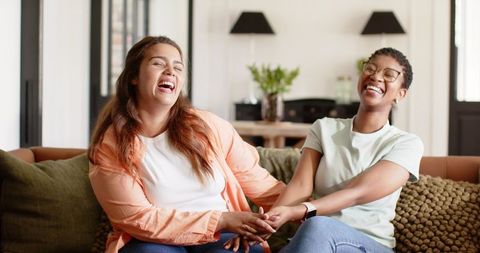 Two african american women laughing and holding hands on cozy living room sofa sharing joy