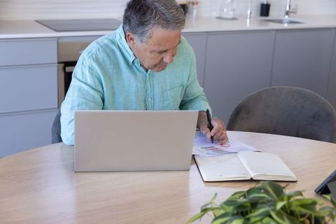 Senior man working from home reviewing financial charts and budget papers at kitchen table