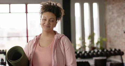 Smiling woman holding rolled yoga mat in bright loft gym wearing pink activewear