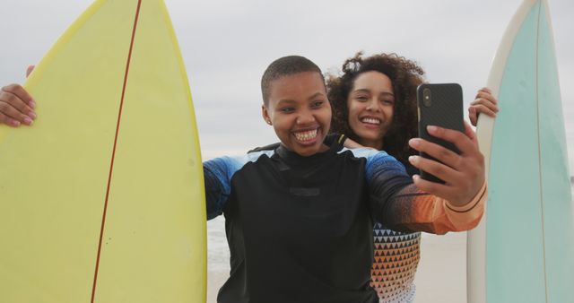 Happy african american female friends on the beach holding surfboards taking selfie. healthy outdoor leisure time by the sea.