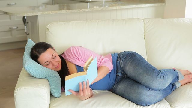 Pregnant woman reading book comfortably on sofa, enjoying quiet time. Ideal for themes like pregnancy, relaxation, leisure, home comfort, casual living.