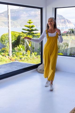 Mature woman enjoying coastal view from modern bright living room wearing mustard jumpsuit