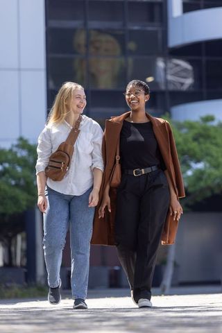 Diverse women laughing and walking in urban plaza carrying brown bags and stylish coats daylight