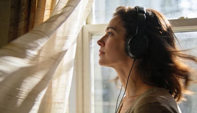 Woman listening to music with over-ear headphones by sunlit window, billowing curtain