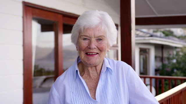 A joyful senior woman stands on her balcony at home, enjoying a moment of relaxation. Her casual attire suggests comfort and ease in her daily routine. This image is perfect for representing themes of active aging, retirement, or home life, and is ideal for use by care agencies, retirement communities, or lifestyle blogs focused on positive living in later years.