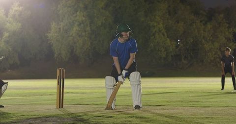 Night cricket batsman preparing to face delivery under floodlights on grassy pitch