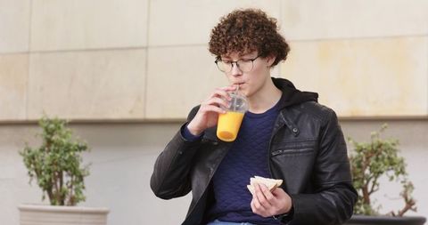 Young man sipping orange juice and eating sandwich on urban bench wearing leather jacket
