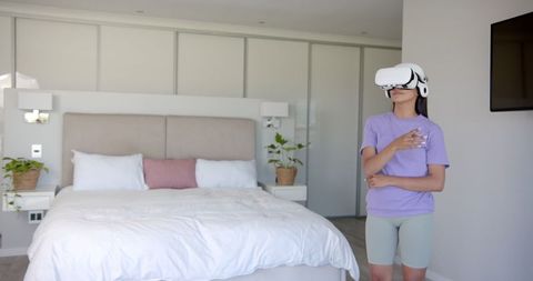 A woman wearing a VR headset stands in a modern bedroom, fully immersed in a virtual world. The room features a minimalist design and neutral tones, providing a contemporary backdrop. This image is ideal for showcasing products or services related to virtual reality, technology advances, or home entertainment. It can also be useful for articles or advertisements focusing on innovative solutions and digital experiences in everyday environments.