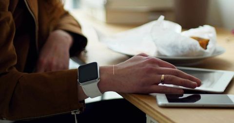 Touching tablet while wearing white smartwatch and gold ring on wooden cafe table