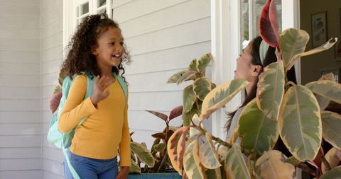 Smiling schoolchild waving at doorway with mint backpack and sunlit porch greenery