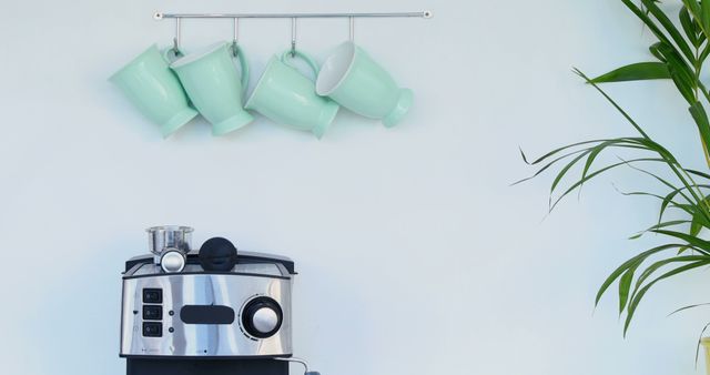 Mint green coffee mugs hang above a modern espresso machine in a clean kitchen setting, with copy space. The minimalist design and pastel colors suggest a contemporary home with a focus on style and simplicity.