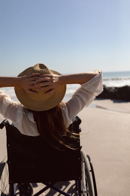 This image shows a disabled woman in a wheelchair enjoying a sunny day at the beach. She is wearing a hat and has her hands behind her head, indicating relaxation and contentment. The ocean waves and clear sky create a serene atmosphere. This image can be used for promoting accessibility, travel, and leisure activities for people with disabilities, as well as for illustrating themes of freedom, independence, and enjoying nature.
