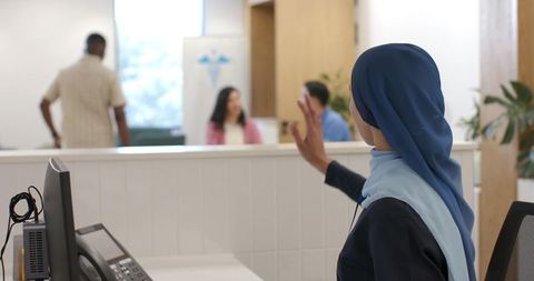 Clinic receptionist wearing blue headscarf waving from modern medical reception desk